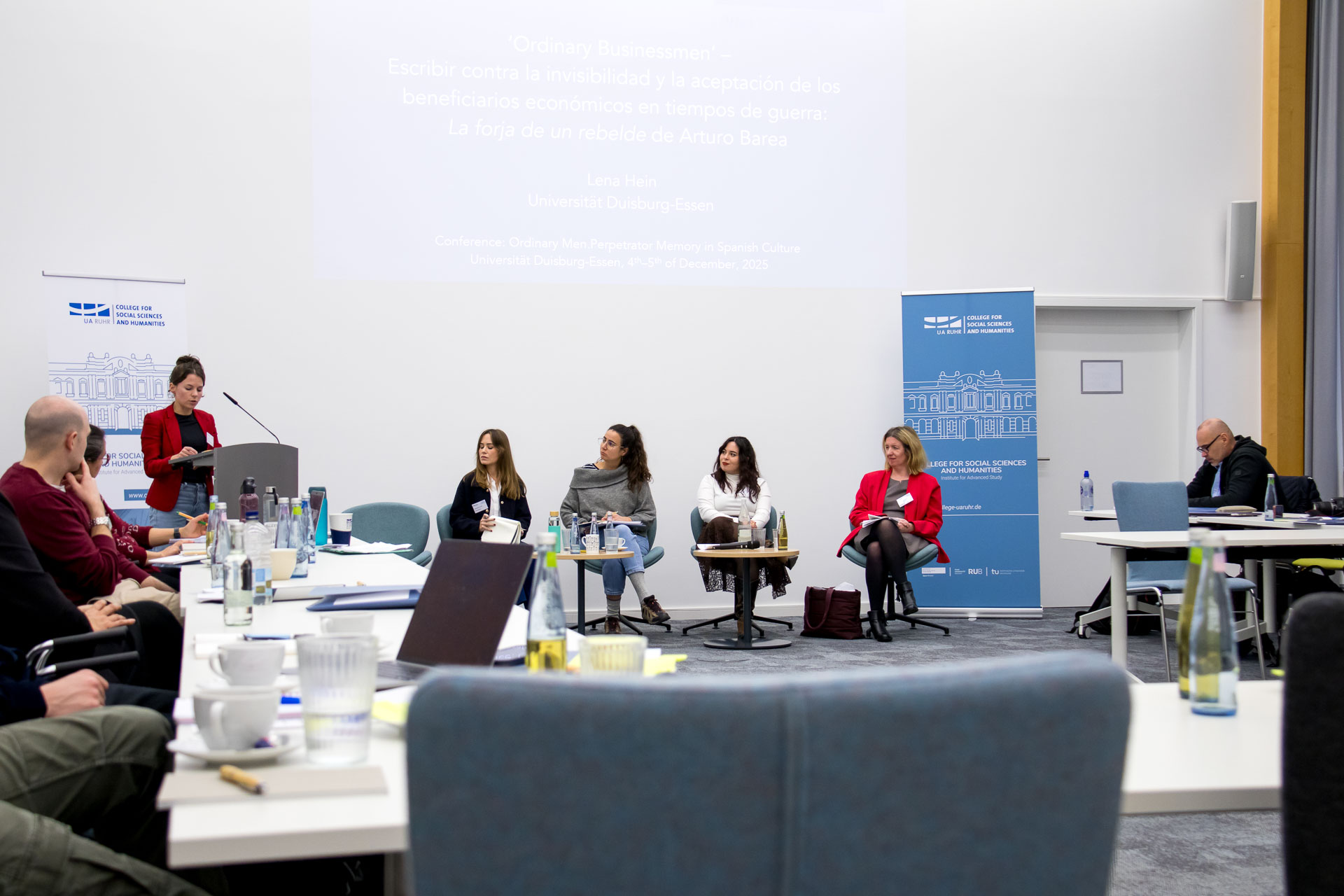 photo from the workshop 'Perpetrator Memory in Spanish culture', participants sitting around a table, a speaker presenting, a screen with slides in the back